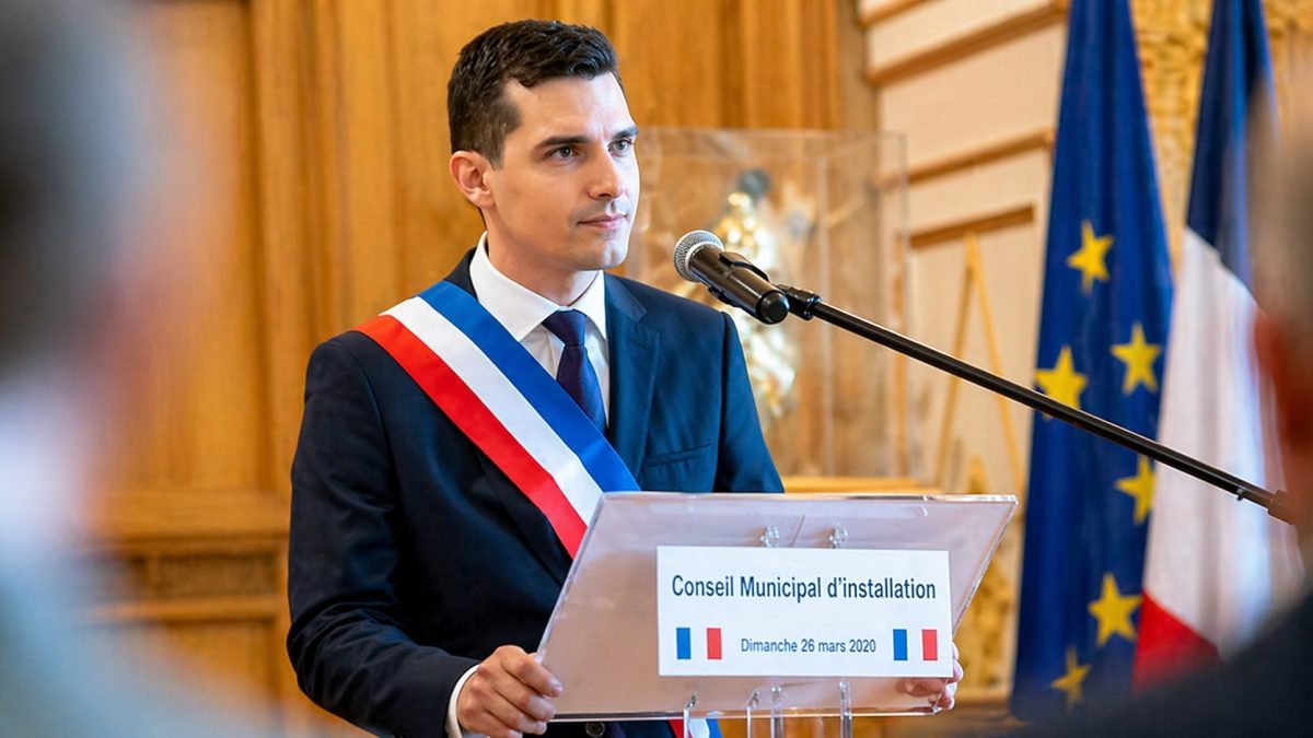Homme politique portant une écharpe tricolore prononçant un discours derrière un pupitre lors d’un conseil municipal, dans une salle officielle avec drapeaux en arrière-plan