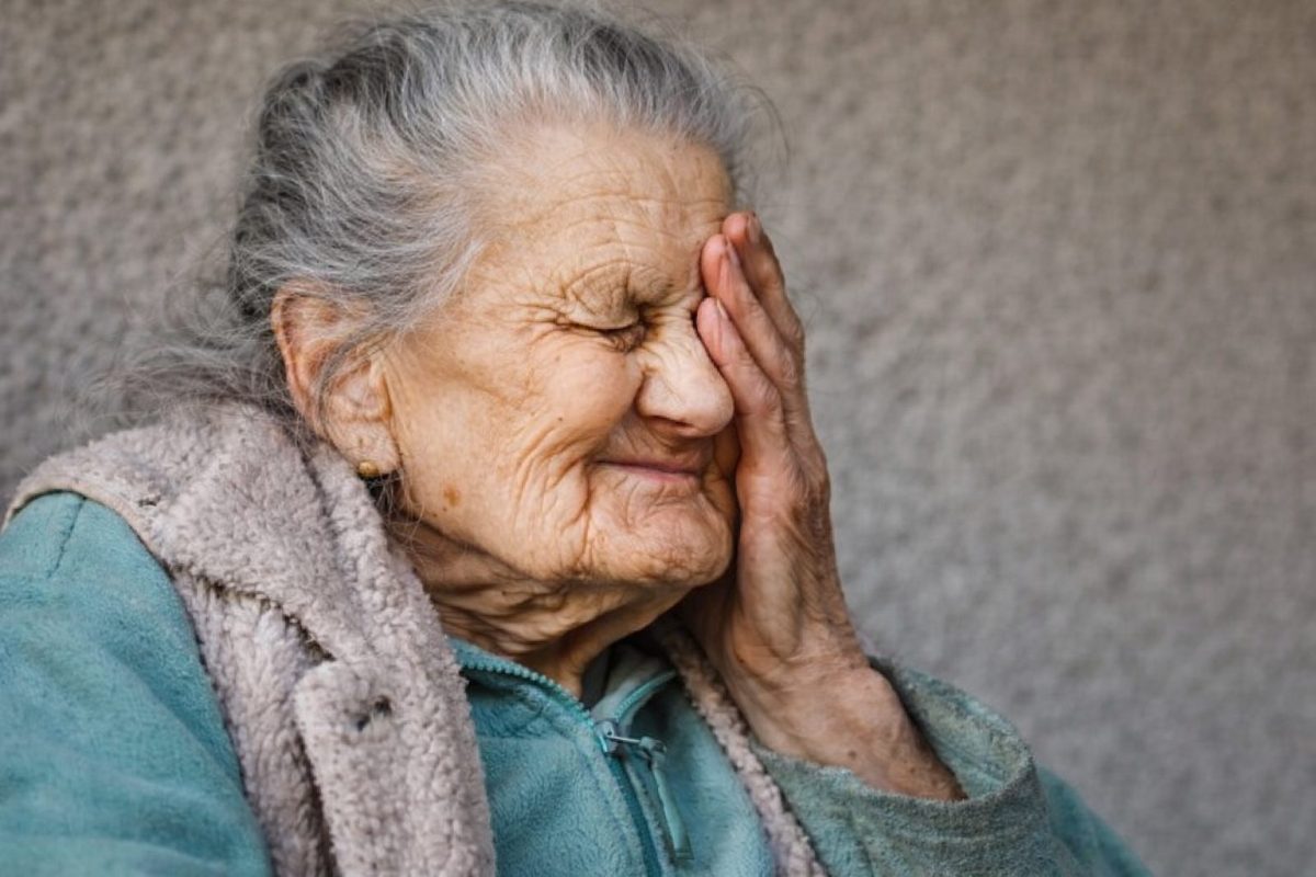 Femme âgée aux cheveux gris, visage marqué par le temps, se tenant la main contre le visage avec une expression mêlant fatigue et agacement, assise devant un mur neutre dans une ambiance calme et réaliste