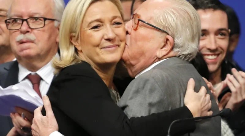 Femme politique blonde souriante enlacée par un homme âgé lors d’un événement public, entourés de personnes applaudissant en arrière-plan, ambiance politique et médiatique