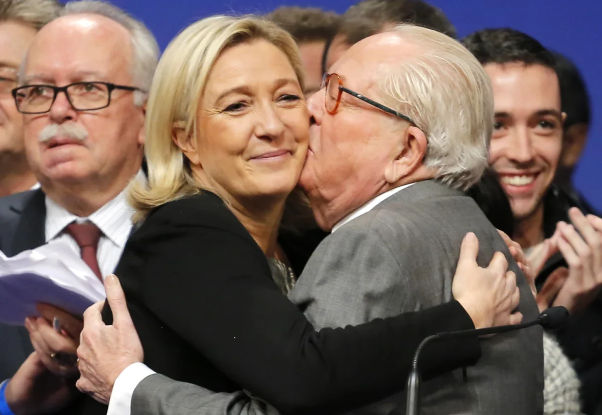 Femme politique blonde souriante enlacée par un homme âgé lors d’un événement public, entourés de personnes applaudissant en arrière-plan, ambiance politique et médiatique