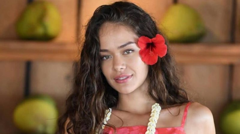 Portrait d’une jeune femme aux cheveux longs et ondulés portant une fleur rouge derrière l’oreille, regard doux et léger sourire, dans un décor intérieur chaleureux avec éléments naturels en arrière-plan