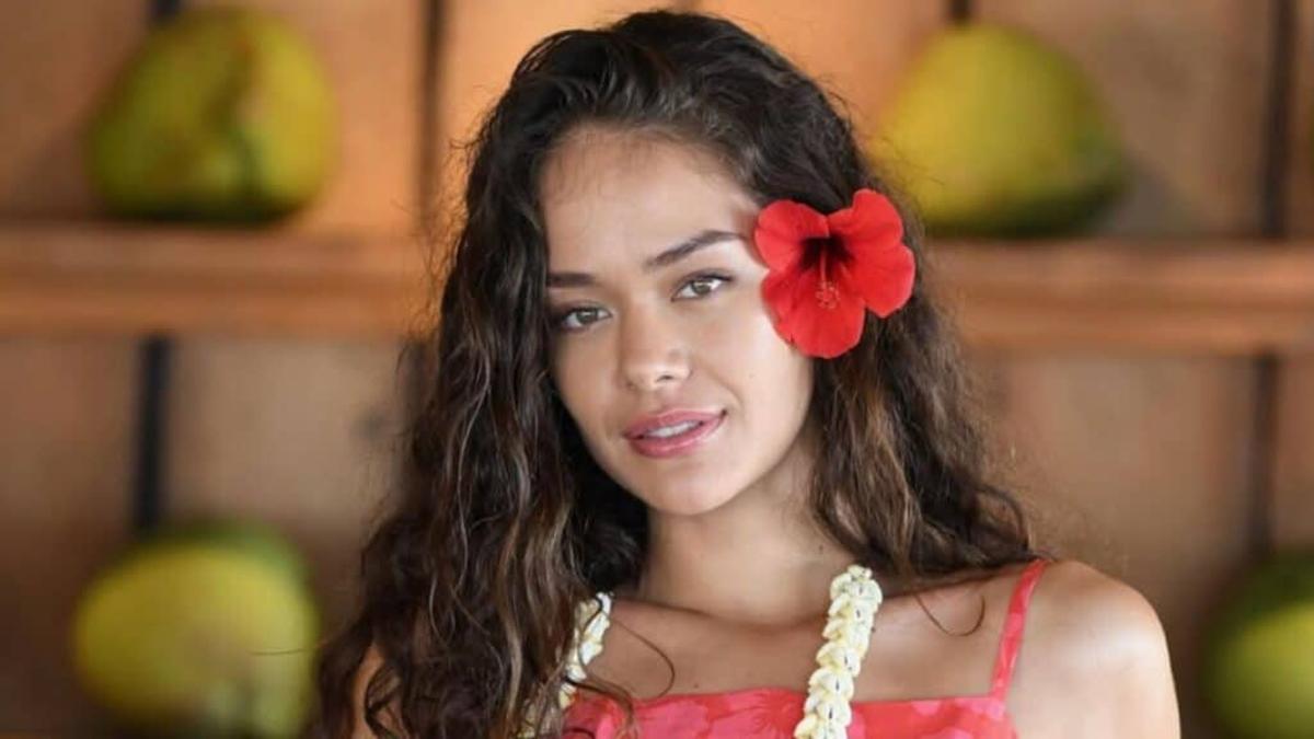 Portrait d’une jeune femme aux cheveux longs et ondulés portant une fleur rouge derrière l’oreille, regard doux et léger sourire, dans un décor intérieur chaleureux avec éléments naturels en arrière-plan