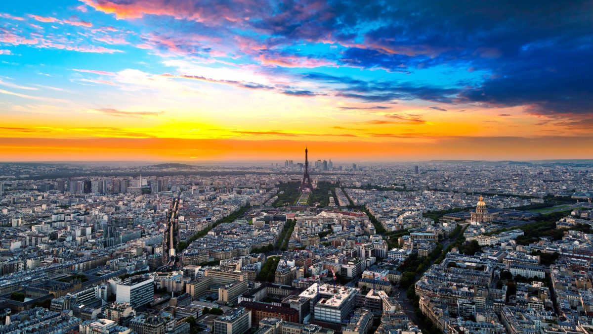 Vue aérienne panoramique d’une grande capitale européenne au coucher du soleil, avec une tour emblématique au centre, des immeubles haussmanniens à perte de vue et un ciel coloré aux nuances orange, rose et bleu