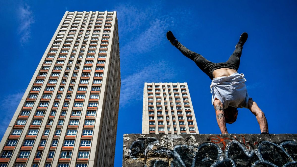 Athlète réalisant une figure acrobatique de parkour en équilibre sur les mains au sommet d’un mur urbain, avec de grands immeubles en arrière-plan sous un ciel bleu dégagé