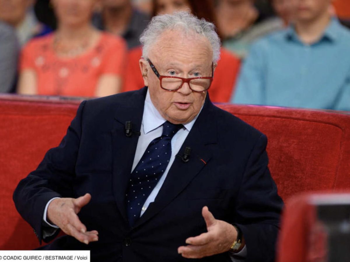 Homme âgé aux cheveux blancs portant des lunettes rouges, assis sur un canapé rouge lors d’une émission télévisée, en train de s’exprimer avec des gestes des mains, public flou en arrière-plan, ambiance de débat ou d’interview