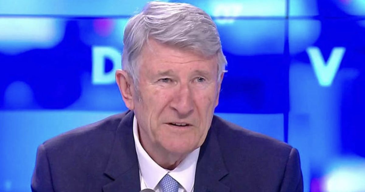 Homme politique invité sur un plateau télévisé en direct, visage sérieux et légèrement ému, éclairage studio bleu en arrière-plan lors d’une émission d’actualité