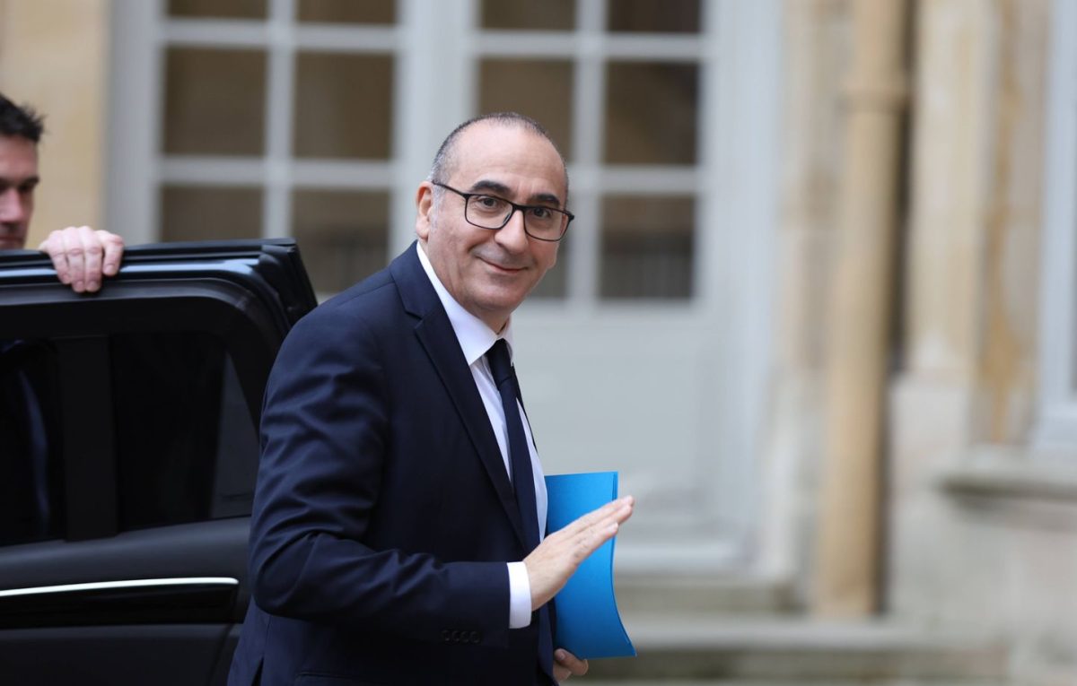 Homme politique en costume saluant en sortant d’un véhicule officiel devant un bâtiment administratif, tenant un dossier bleu, dans un contexte lié à la réforme des délais de traitement des titres de séjour en France