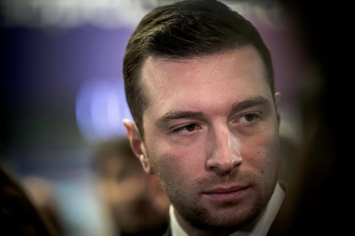 Portrait d’un homme politique au regard sérieux lors d’un événement public, entouré de personnes en arrière-plan flou