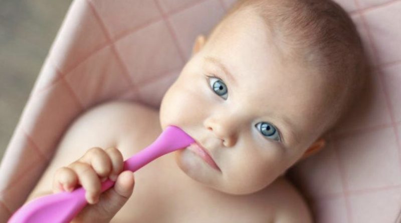 Bébé aux yeux clairs allongé dans un transat, tenant une cuillère rose, regard expressif et curieux, illustrant un prénom féminin d’origine arabe tendance en France