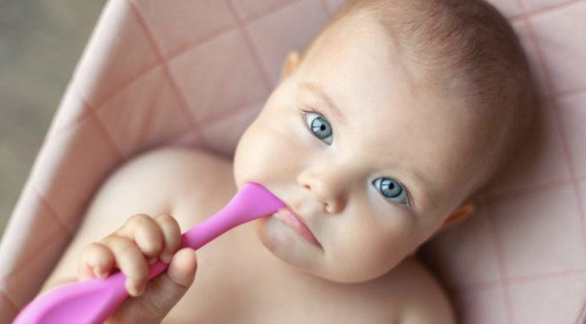 Bébé aux yeux clairs allongé dans un transat, tenant une cuillère rose, regard expressif et curieux, illustrant un prénom féminin d’origine arabe tendance en France