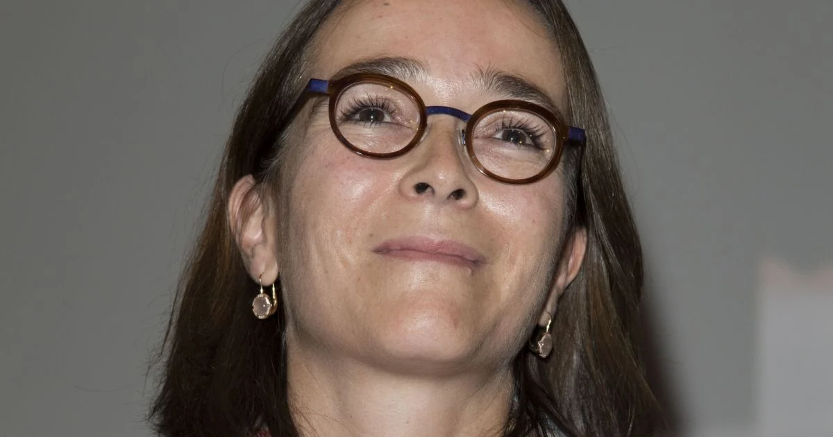 Portrait en gros plan d’une femme dirigeante du secteur audiovisuel portant des lunettes rondes, visage légèrement levé avec un léger sourire, éclairage neutre sur fond flou, illustrant une personnalité influente au cœur d’une polémique liée à la gestion et à la rémunération dans le service public audiovisuel.