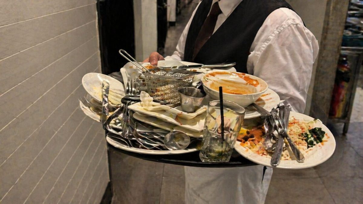 Serveur transportant un plateau surchargé de vaisselle sale avec assiettes empilées, couverts en désordre et restes de nourriture dans un couloir de restaurant, illustrant les erreurs fréquentes des clients qui compliquent le service en salle