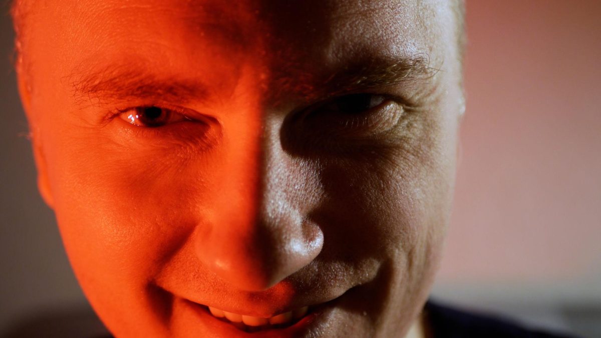 Visage d’une personne en gros plan avec un sourire ambigu et un regard intense, éclairé par une lumière rouge et sombre, illustrant une apparence rassurante dissimulant une personnalité manipulatrice
