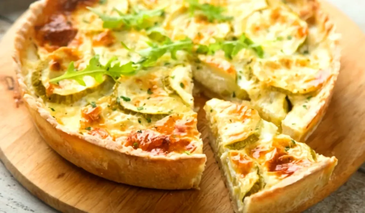 Tarte aux légumes cuite au four avec une pâte brisée dorée et fondante, garnie de rondelles de courgettes, fromage gratiné et herbes fraîches, présentée sur une planche en bois