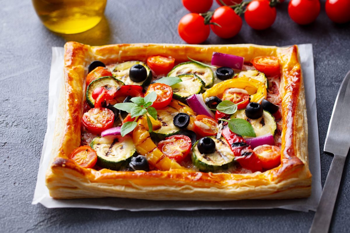 Tarte aux légumes cuite avec une pâte feuilletée croustillante et dorée, garnie de tomates cerises, courgettes, olives noires, poivrons et oignons rouges, avec une texture feuilletée bien visible sur les bords