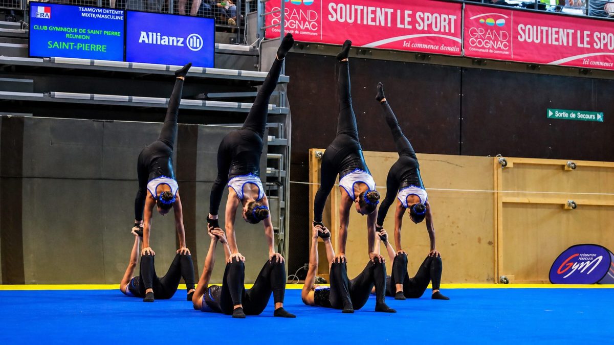 Équipe de gymnastes en TeamGym réalisant une figure acrobatique synchronisée en équilibre sur les mains lors d’une compétition sur tapis bleu
