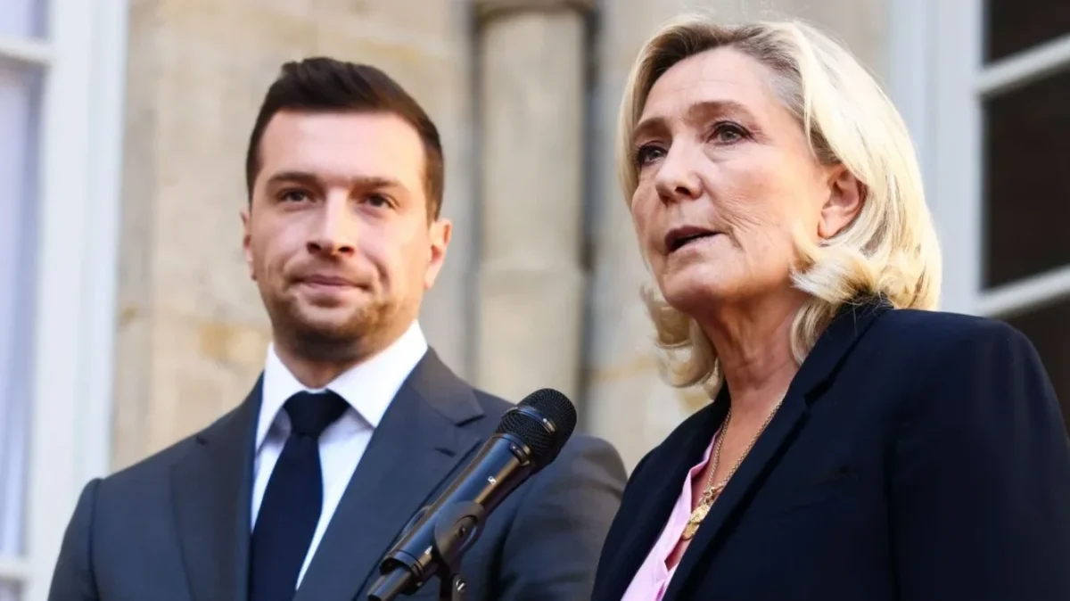 Deux responsables politiques français debout côte à côte lors d’une prise de parole publique en extérieur, l’un en costume sombre regardant attentivement, l’autre s’exprimant au micro avec une expression sérieuse, illustrant un contexte de tensions politiques et d’enjeux électoraux.