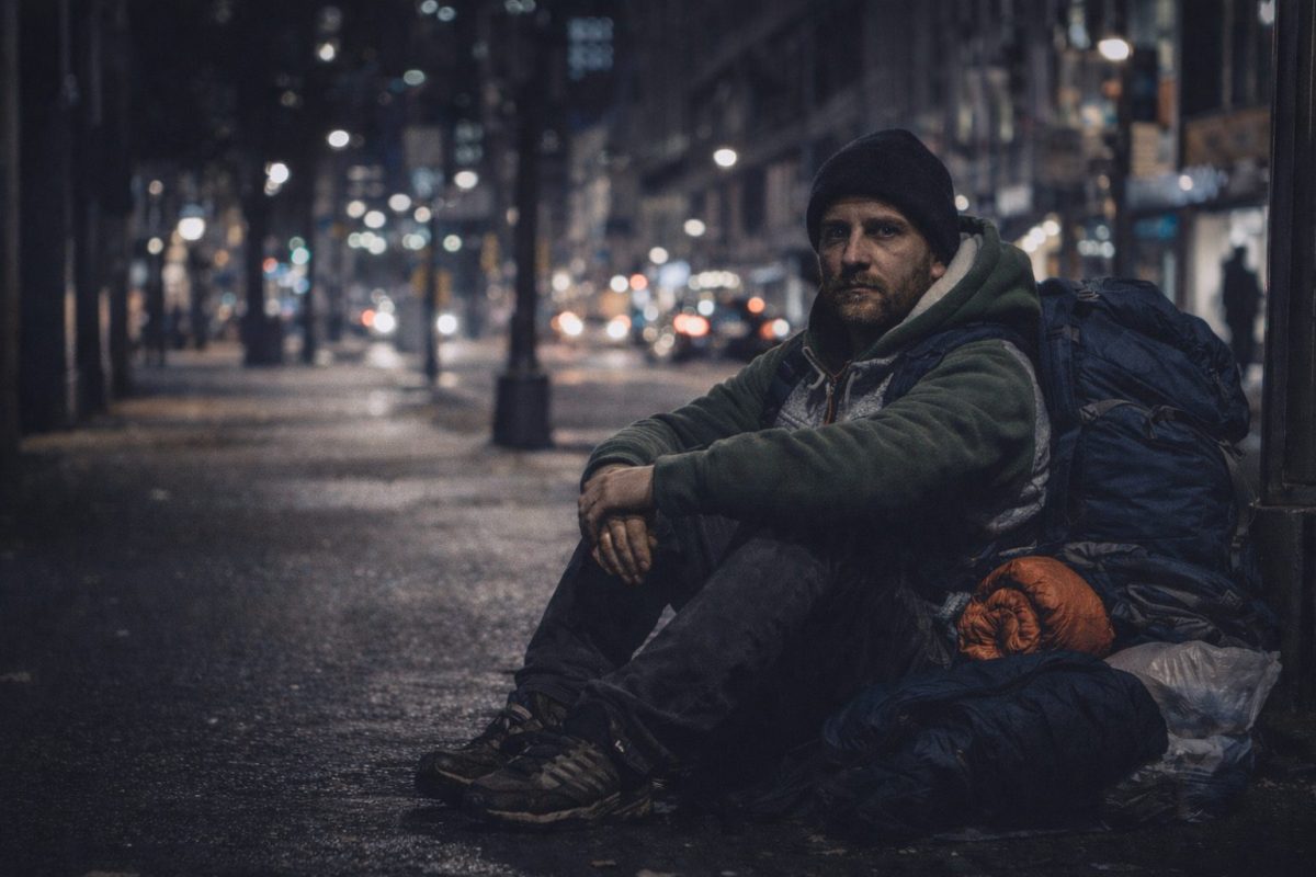 Homme seul assis sur un trottoir de ville la nuit avec un sac à dos et des affaires personnelles, regard fatigué mais déterminé, scène urbaine sombre illustrant la précarité d’un travailleur sans logement malgré un emploi
