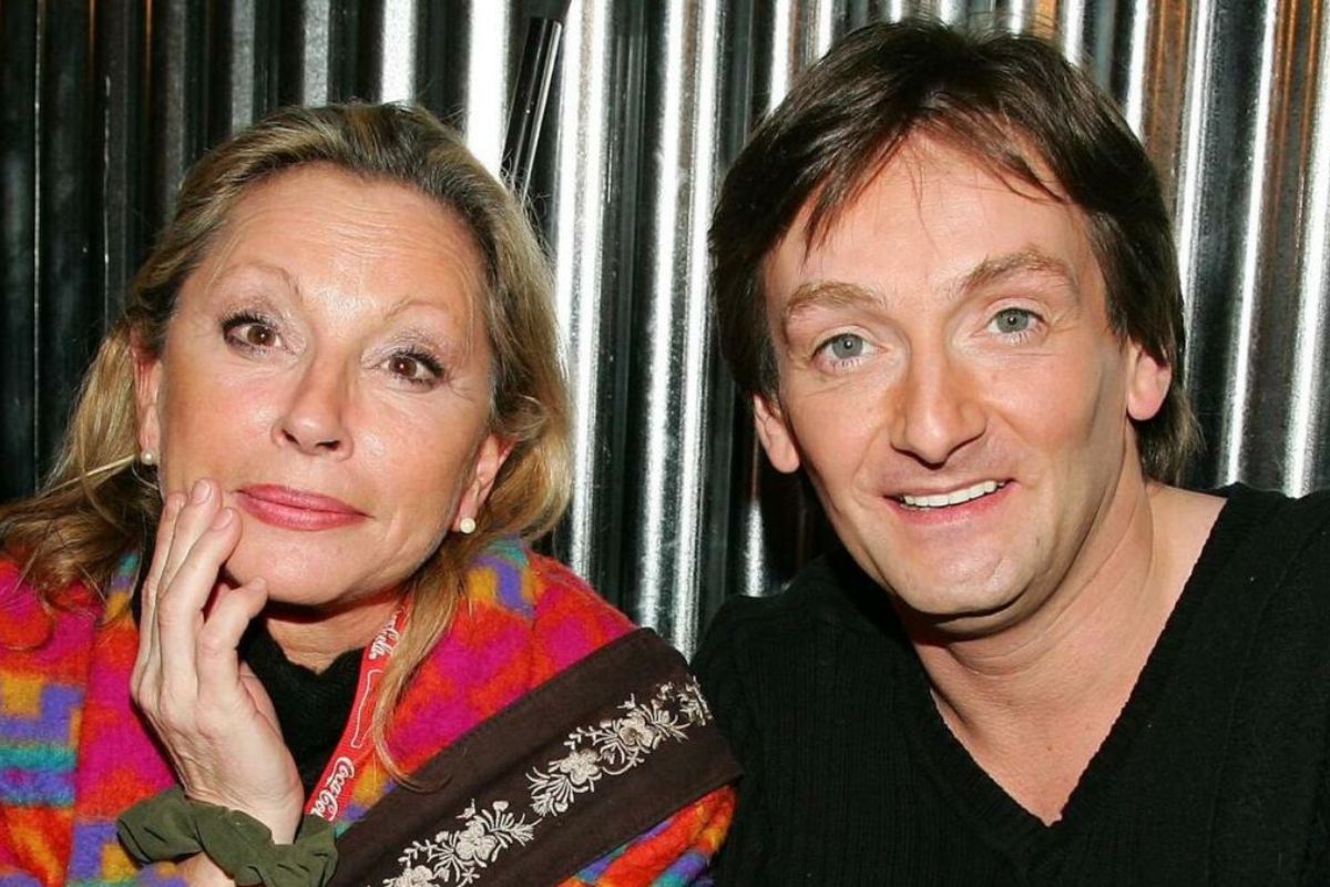 Couple de célébrités posant côte à côte, souriants face à l’objectif, la femme appuie son visage sur sa main avec une expression douce tandis que l’homme regarde l’appareil avec assurance, illustrant une relation médiatisée des années 90