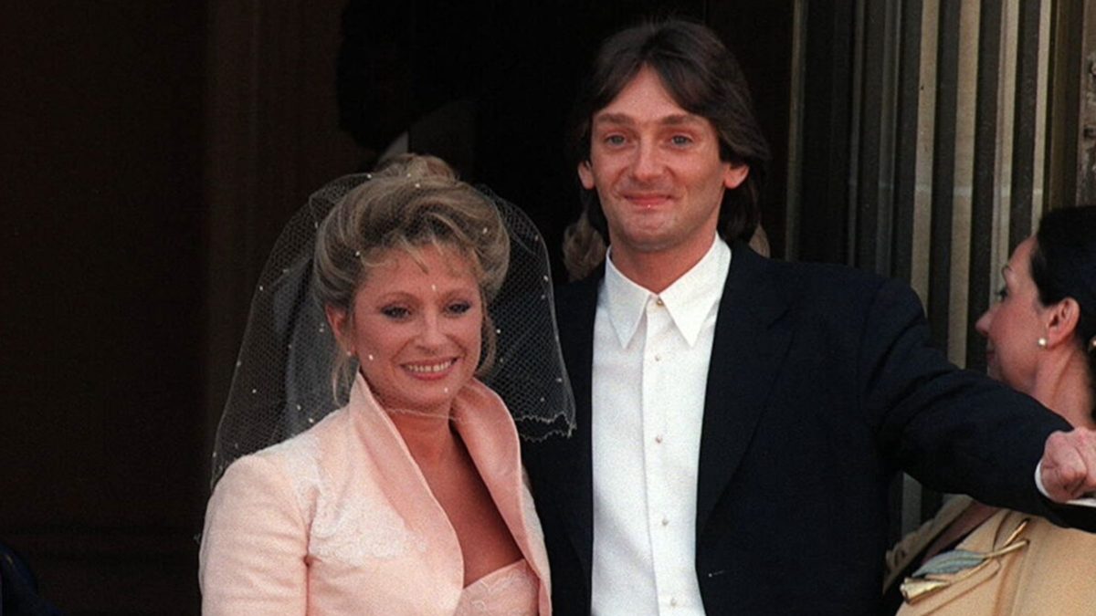 Couple de célébrités lors d’un mariage dans les années 90, la mariée porte un voile et un tailleur clair tandis que le marié en costume sombre sourit face aux invités, scène symbolisant une union médiatisée et une histoire d’amour atypique sous le regard du public
