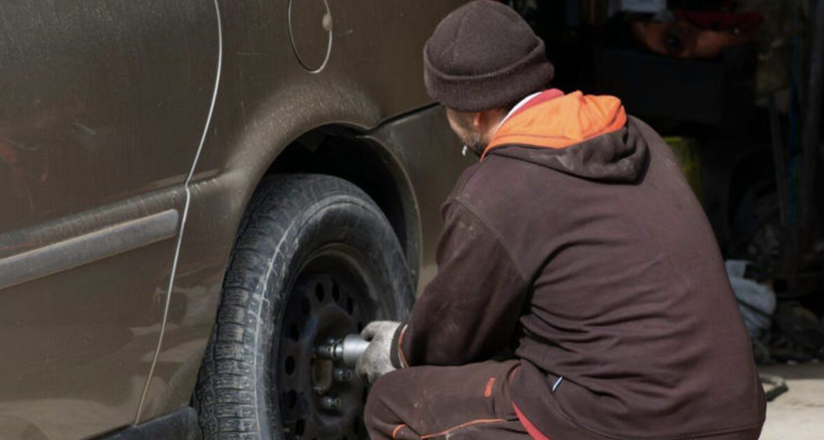 Individu accroupi en train de démonter la roue d’une voiture à l’aide d’une clé, portant des gants et un bonnet, concentré sur l’écrou du pneu, scène capturée de côté dans un environnement de type garage ou extérieur, illustrant une situation de retrait ou de vol de roues sur un véhicule, avec une ambiance réaliste et discrète liée à un fait divers automobile.