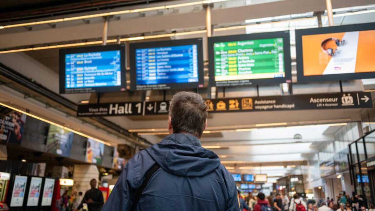 Voyageur de dos observant les panneaux d’affichage des départs dans une grande gare moderne européenne avant de monter dans un train, dans un contexte lié à une amende reçue après l’achat d’un billet quelques secondes trop tard.
