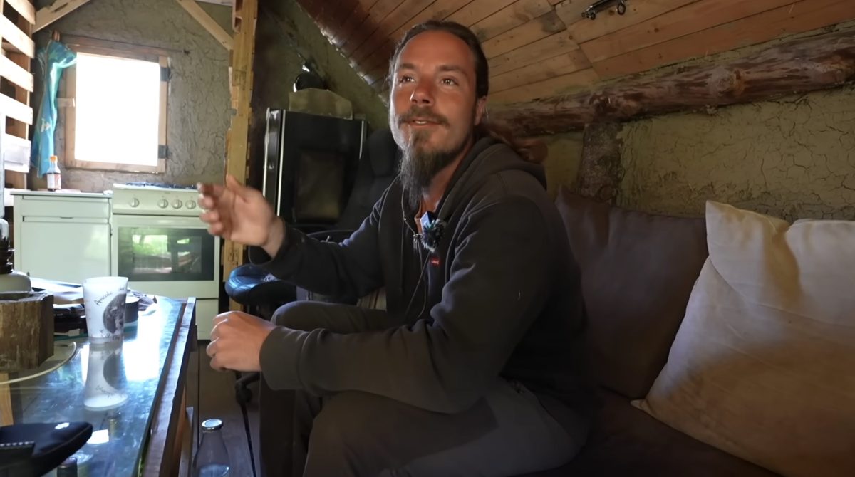 Antonin est assis à l’intérieur de sa maison auto-construite dans le Puy-de-Dôme. Entouré de matériaux naturels, de mobilier simple et d’équipements du quotidien, il explique son mode de vie basé sur l’autonomie, la récupération, la sobriété volontaire et la réduction des dépenses.