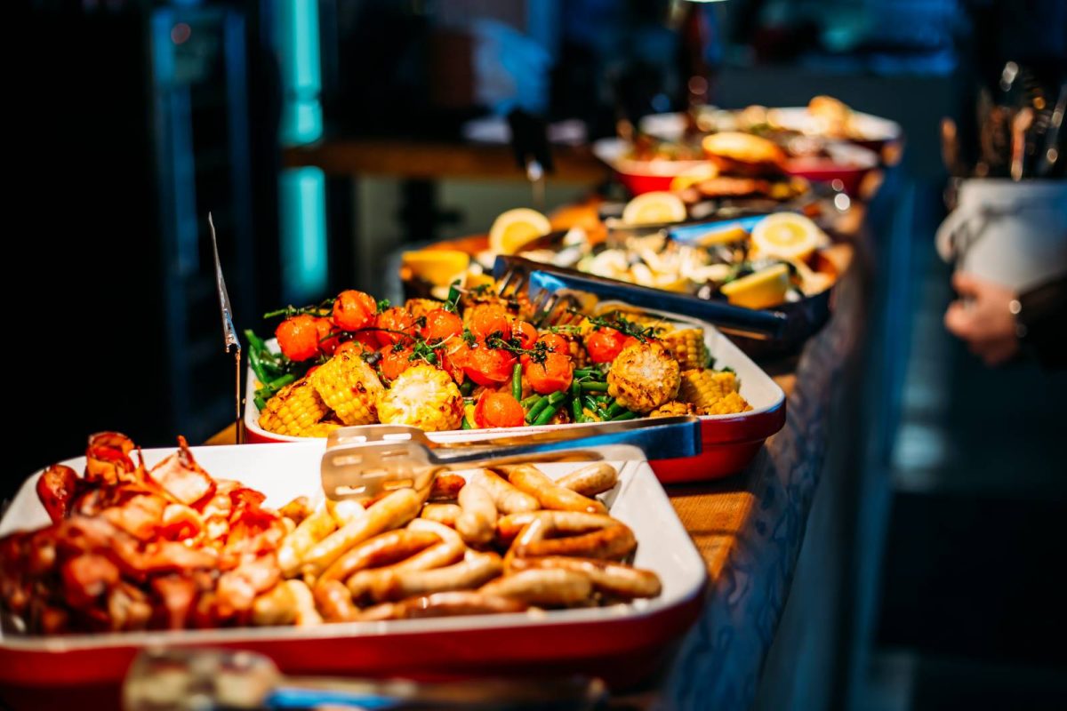 Buffet à volonté avec plusieurs plats chauds abondants disposés sur une longue table de restaurant, légumes grillés, saucisses et plats servis en libre-service dans une ambiance chaleureuse, illustration du gaspillage alimentaire et des nouvelles règles dans certains restaurants à volonté.