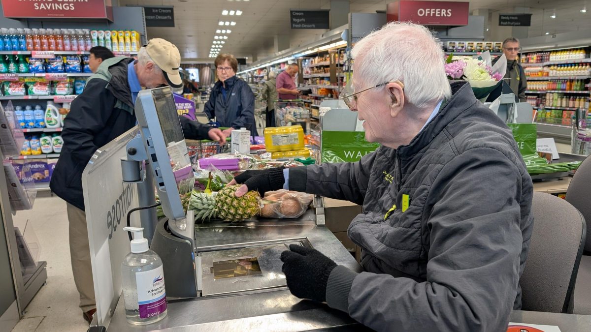 un homme âgé travaille comme caissier dans un supermarché américain en scannant les courses de clients dans une grande surface moderne avec des rayons alimentaires visibles à l’arrière-plan
