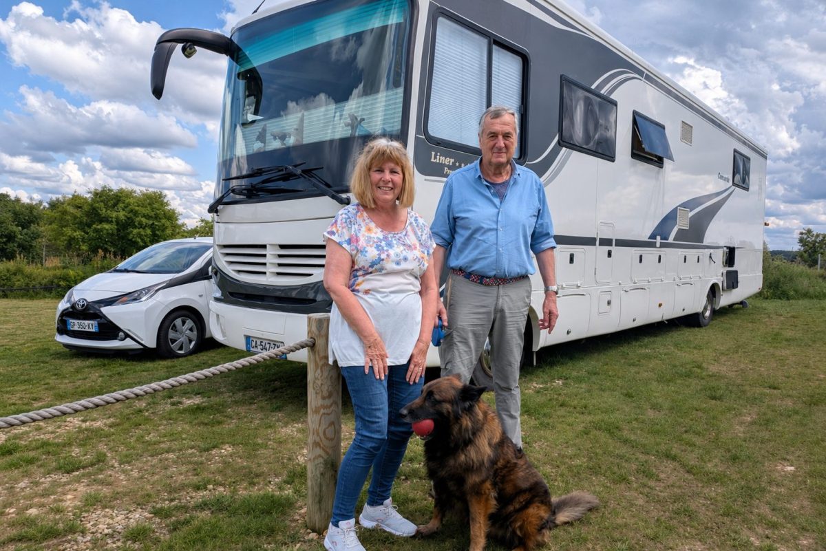 Couple de retraités posant devant un immense camping-car blanc haut de gamme stationné sur une aire herbeuse en Bretagne, accompagné de leur chien et d’une petite voiture, sous un ciel nuageux.