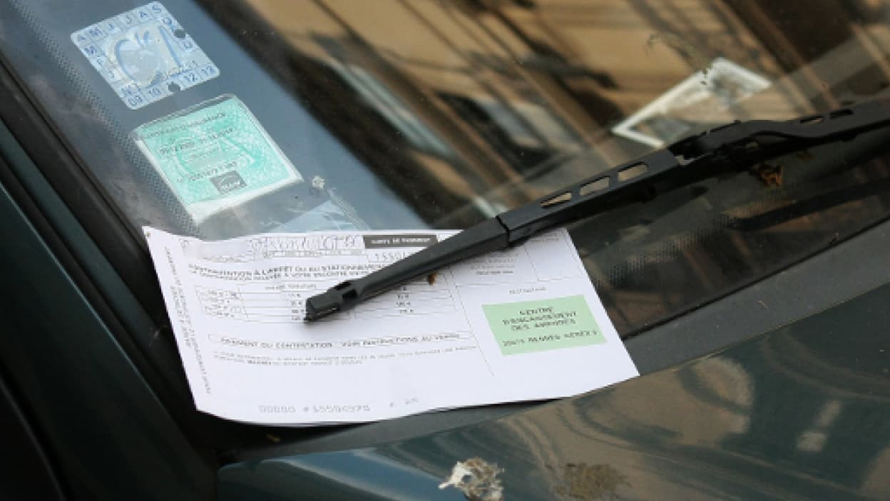 Contravention glissée sous l’essuie-glace d’une voiture stationnée en ville avec vignette visible sur le pare-brise, illustrant une amende de stationnement et les sanctions liées au stationnement gênant.