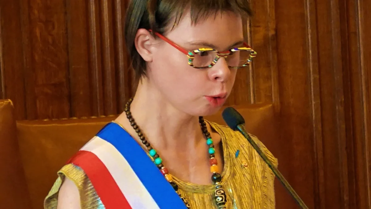Éléonore Laloux prend la parole derrière un micro avec son écharpe tricolore de conseillère municipale à Arras, symbole de son engagement pour l’inclusion des personnes porteuses de trisomie 21 dans la vie politique française