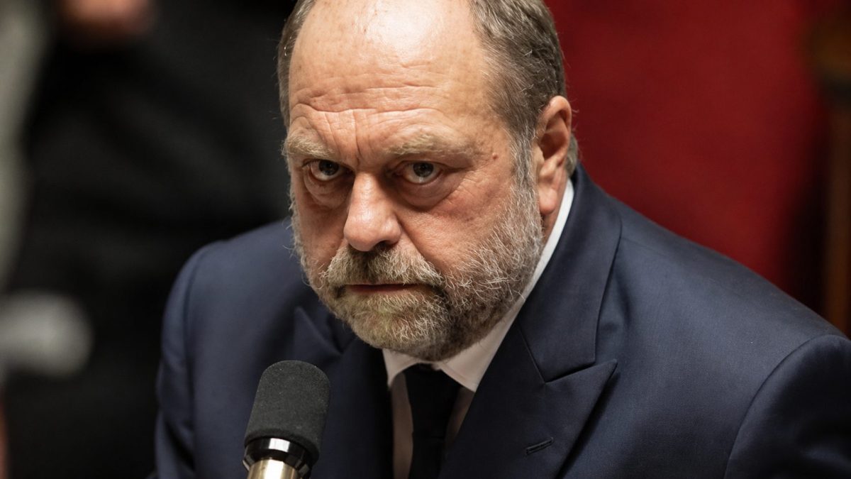 Gros plan sur un homme politique français barbu en costume sombre, assis devant un microphone avec un regard sérieux et tendu dans une ambiance médiatique et institutionnelle.