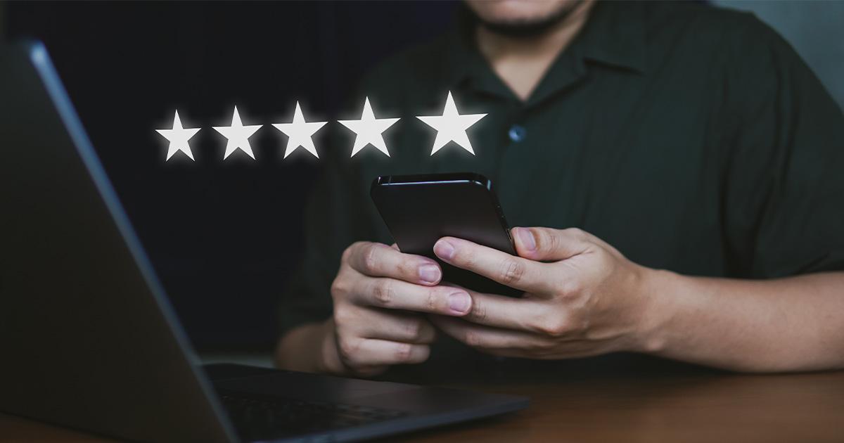 personne utilisant un smartphone devant un ordinateur portable avec des étoiles de notation cinq étoiles affichées à l’écran, illustration des avis Google et de la réputation en ligne des entreprises