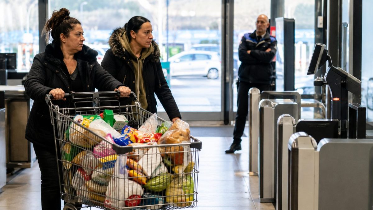 Deux femmes poussant un caddie rempli dans un supermarché moderne près des caisses automatiques, pendant qu’un agent de sécurité les observe discrètement à distance.