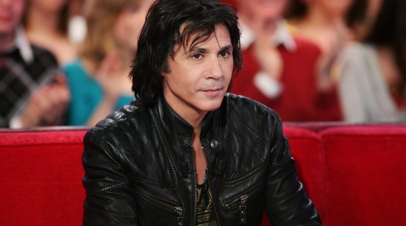 Jean-Luc Lahaye assis sur un canapé rouge lors d’une émission télévisée, portant une veste noire en cuir, dans un contexte de polémique autour de ses concerts programmés dans des mairies RN malgré les accusations de violences sexuelles visant le chanteur.