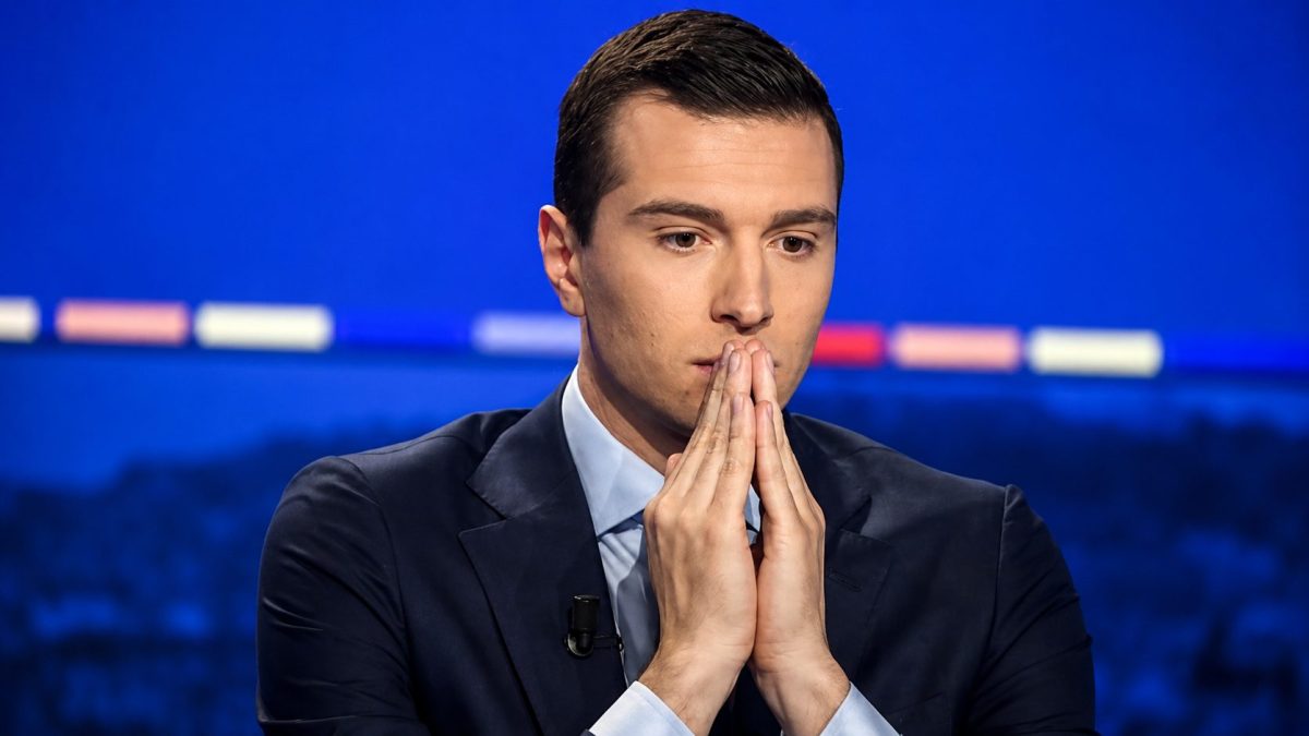 Portrait d’un jeune responsable politique français en costume sombre sur un plateau télévisé bleu, les mains jointes devant le visage avec une expression grave et réfléchie.