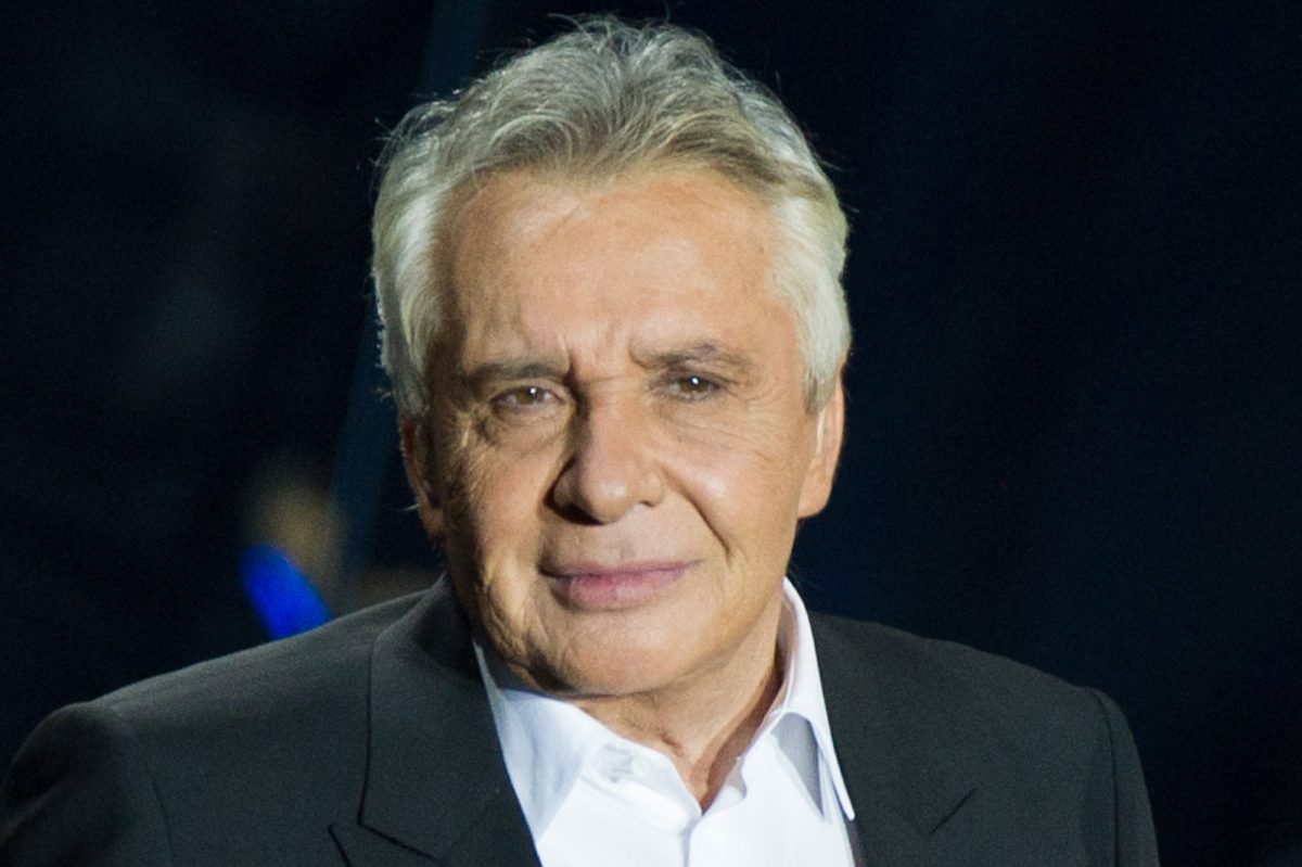 Michel Sardou lors d’une apparition publique, vêtu d’une veste sombre et d’une chemise claire, regardant l’objectif. Le chanteur français est au cœur de l’actualité après avoir apporté son soutien à Kylian Mbappé concernant ses prises de position sur le Rassemblement National.