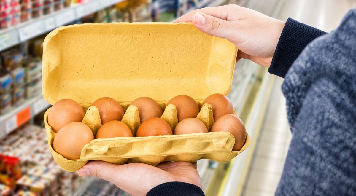 Personne tenant une boîte d’œufs ouverte dans un supermarché avec plusieurs œufs bruns visibles et un rayon alimentaire flou en arrière-plan, illustrant la vérification de l’origine des œufs vendus en grande surface.