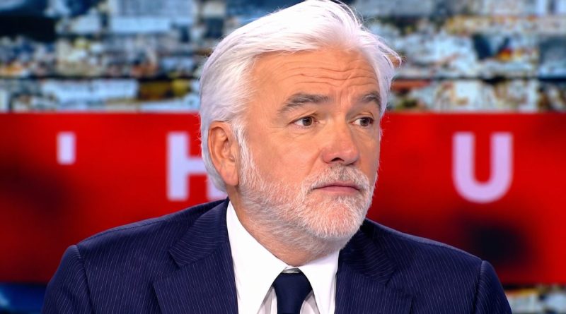 animateur de télévision français en costume sombre sur un plateau d’émission d’actualité avec expression sérieuse et arrière-plan flou rouge et lumineux