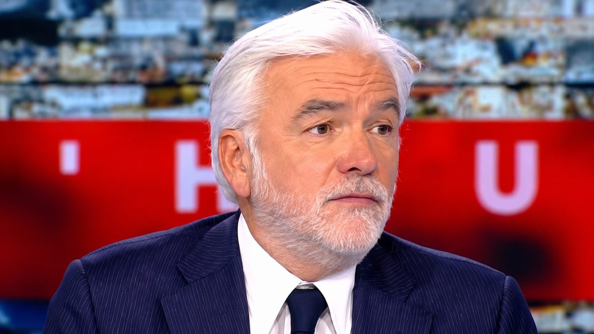 animateur de télévision français en costume sombre sur un plateau d’émission d’actualité avec expression sérieuse et arrière-plan flou rouge et lumineux