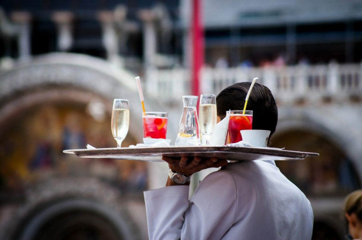 Serveur portant un plateau rempli de verres et de consommations sur une terrasse de restaurant animée, dans une ambiance de service intense en pleine affluence.