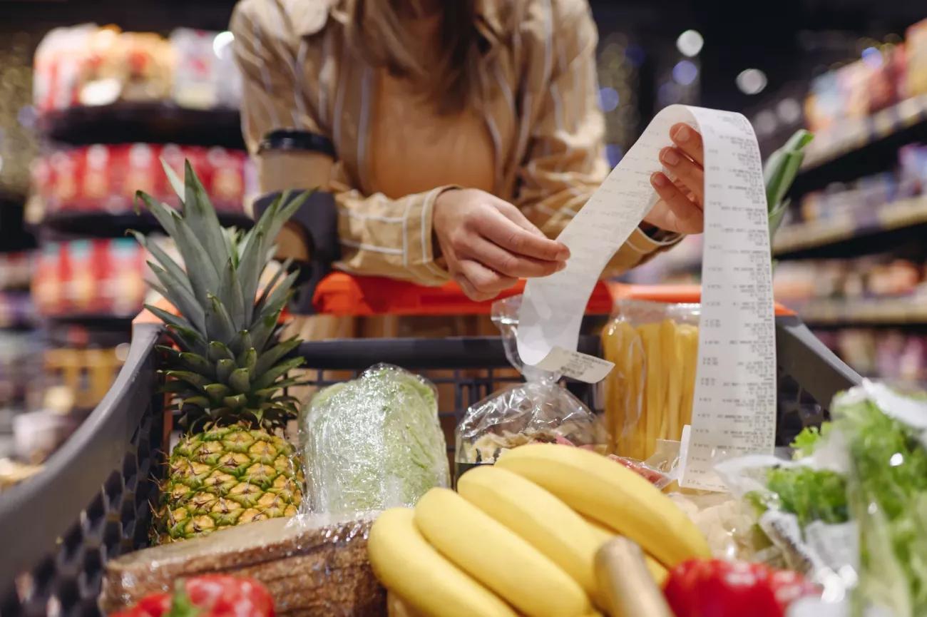 Personne faisant ses courses dans un supermarché avec un long ticket de caisse à la main et un chariot rempli de produits alimentaires, illustrant la hausse du coût de la vie et les difficultés financières des ménages en France.