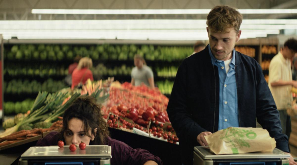 Cliente observant de très près une balance de caisse automatique dans un supermarché pendant qu’un homme pèse un sac de fruits et légumes, illustrant une erreur fréquente de pesée pouvant fausser le prix affiché.
