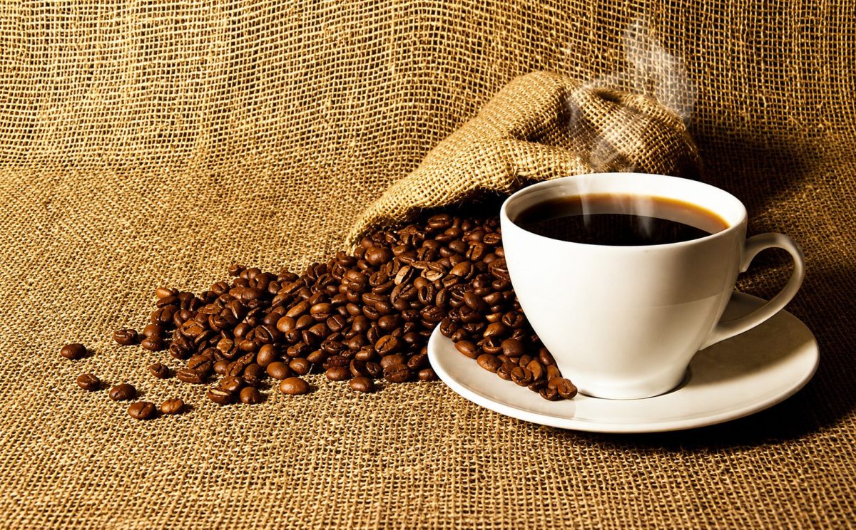 Tasse de café noir fumant posée sur une soucoupe blanche au milieu de grains de café renversés sur une toile en jute, illustration des bienfaits et risques du café et de la caféine sur la santé et le sommeil