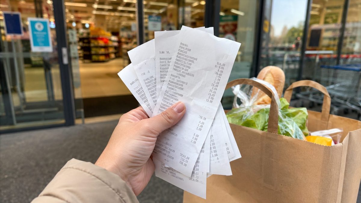 Main tenant plusieurs tickets de caisse en papier thermique à la sortie d’un supermarché moderne, avec des courses alimentaires en arrière-plan et une ambiance réaliste du quotidien.