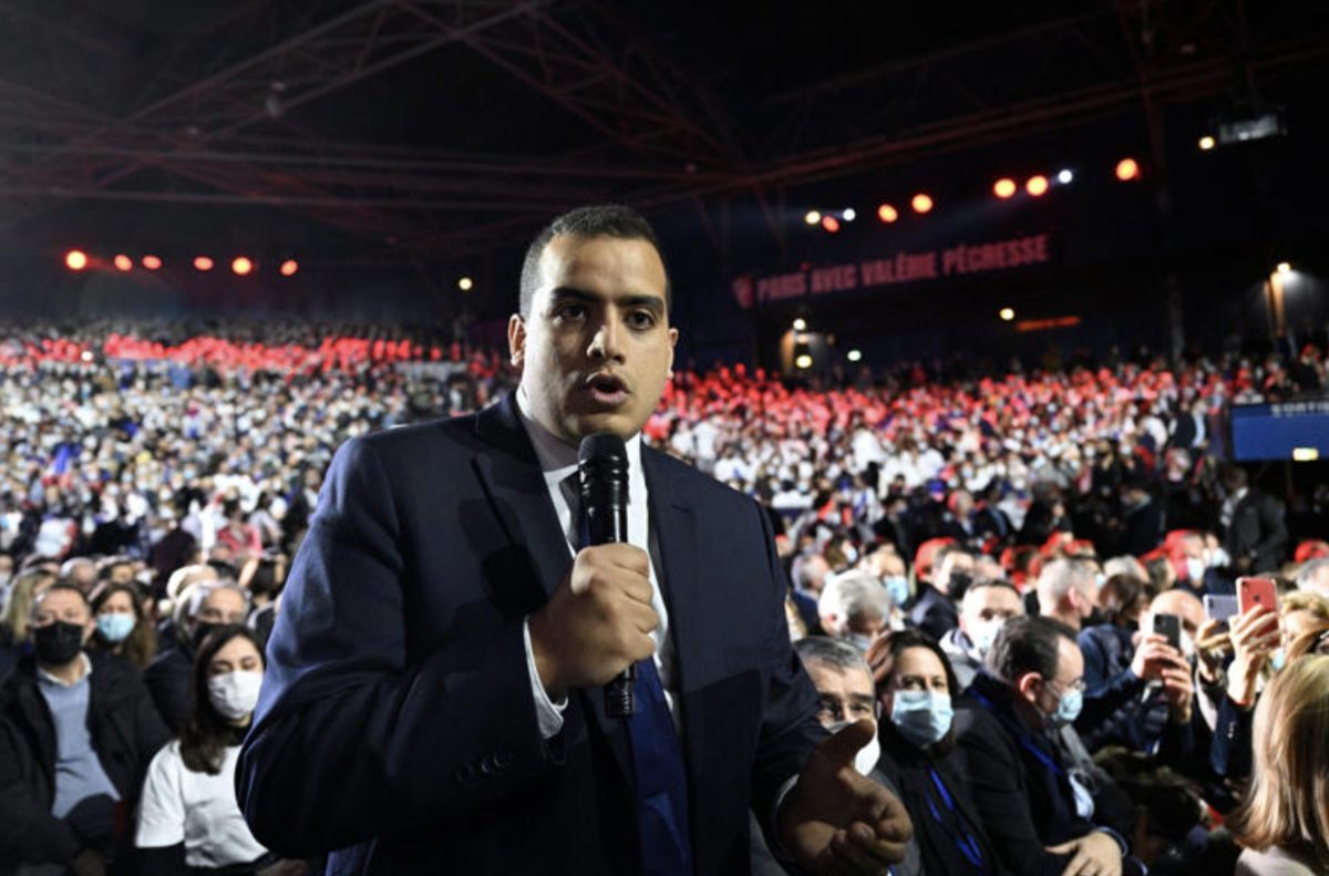 Journaliste tenant un micro au milieu d'une foule nombreuse lors d'un grand rassemblement politique en intérieur, sous les projecteurs d'une vaste salle remplie de participants. L'image illustre un débat médiatique et politique fortement médiatisé dans un contexte de tensions autour du pluralisme et de la liberté d'expression.