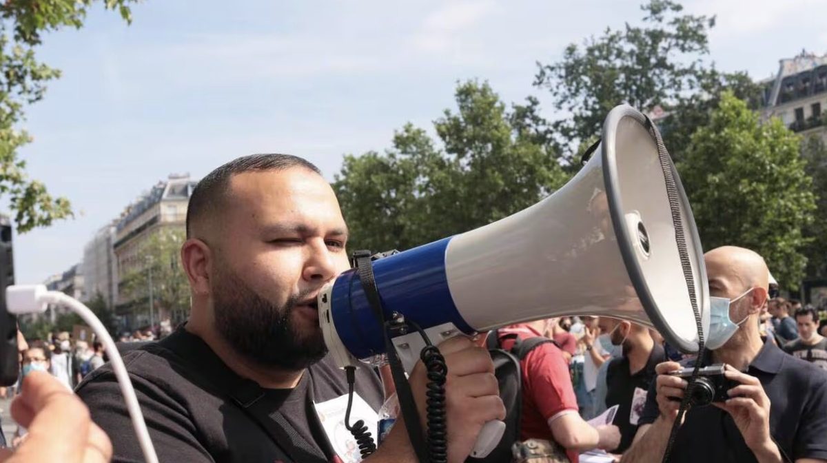 Anasse Kazib s’exprime dans un mégaphone lors d’un rassemblement politique en plein air. Entouré de manifestants et de militants dans une rue urbaine, le syndicaliste et candidat déclaré à la présidentielle 2027 prend la parole devant une foule mobilisée. Cette image illustre son engagement politique, social et militant dans le cadre de sa campagne présidentielle.