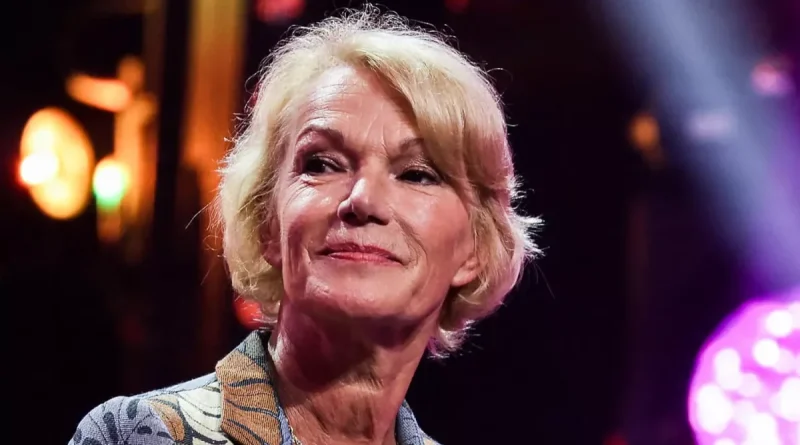 Femme blonde d’environ 70 ans lors d’une apparition publique, regard tourné vers l’horizon, dans une ambiance lumineuse de plateau ou d’événement médiatique. Son expression calme et réfléchie illustre un moment de confidence sur la transmission, les liens affectifs construits au fil du temps et les projets personnels à un âge avancé.