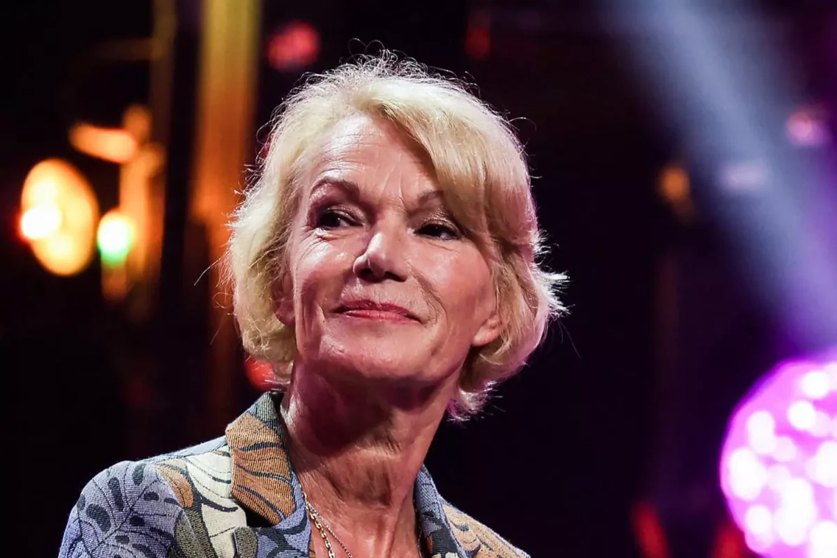 Femme blonde d’environ 70 ans lors d’une apparition publique, regard tourné vers l’horizon, dans une ambiance lumineuse de plateau ou d’événement médiatique. Son expression calme et réfléchie illustre un moment de confidence sur la transmission, les liens affectifs construits au fil du temps et les projets personnels à un âge avancé.